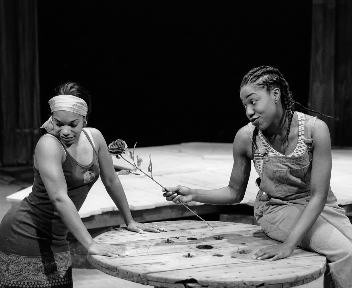 Two performers in a black-and-white stage scene, one offering a rose to the other.